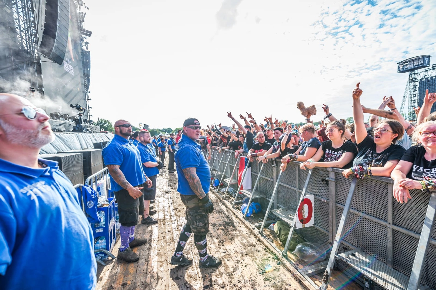 Ronin – Sicherheitsdienst – Sicherheitskräfte in blauen Hemden stehen im abgesperrten Bereich zwischen einer großen Konzertbühne und einer ausgelassenen Festivalmenge.
